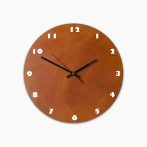 Puede incluir: Un reloj de pared redondo con una esfera marrón texturizada y números blancos. Las manecillas del reloj son negras. El reloj es un artículo decorativo para la decoración del hogar.