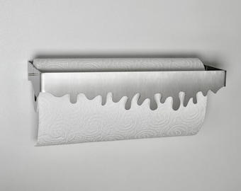 Puede incluir: Un portarrollos de papel de acero inoxidable montado en una pared blanca. El soporte tiene un diseño único con un borde ondulado y goteante. Un rollo de toallas de papel blancas con un patrón de remolino está parcialmente desenrollado.