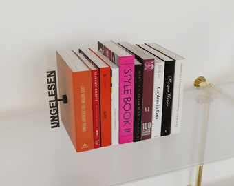 May include: A set of books on a glass shelf with a black metal bookend that says "UNGELESEN". The books are mostly black with white text, but there are also a few red and pink books. The books include titles like "Roger Vivier", "Gardens in Paris", "100 Fashion", "Vanity Fair", "Style Book II", "Alaia", "Yves Saint Laurent", and "Louis Vuitton 100 Legendary Trunks".