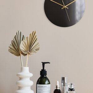 May include: A black clock with gold hands on a beige wall, a white vase with dried palm leaves, and a variety of bottles of lotion and serum on a marble surface. The bottles are labeled "lovgron", "Elie Saab", "EISENBERG", and "L'INTERDIT".