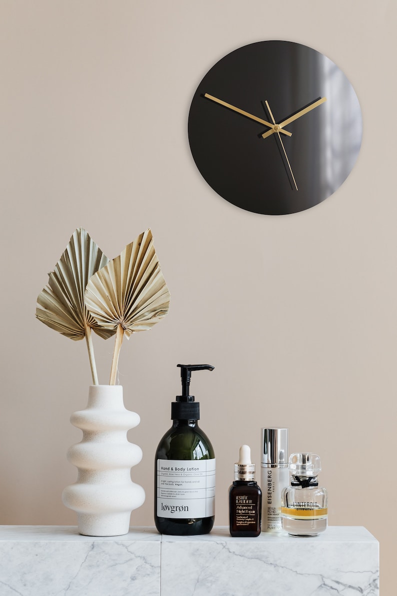 May include: A black clock with gold hands on a beige wall, a white vase with dried palm leaves, and a variety of bottles of lotion and serum on a marble surface. The bottles are labeled "lovgron", "Elie Saab", "EISENBERG", and "L'INTERDIT".