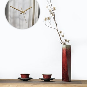 May include: A wooden table with two red teacups and saucers, a red vase with a dried flower arrangement, and a round silver clock on the wall behind the table.