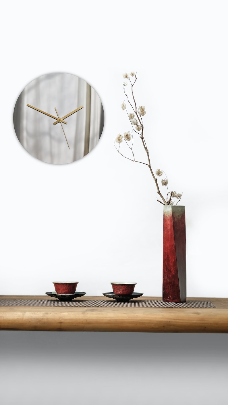May include: A wooden table with two red teacups and saucers, a red vase with a dried flower arrangement, and a round silver clock on the wall behind the table.