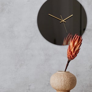 May include: A black round clock with gold hands hangs on a gray wall. A beige ceramic vase with a round body and a smaller round base holds a dried orange flower.