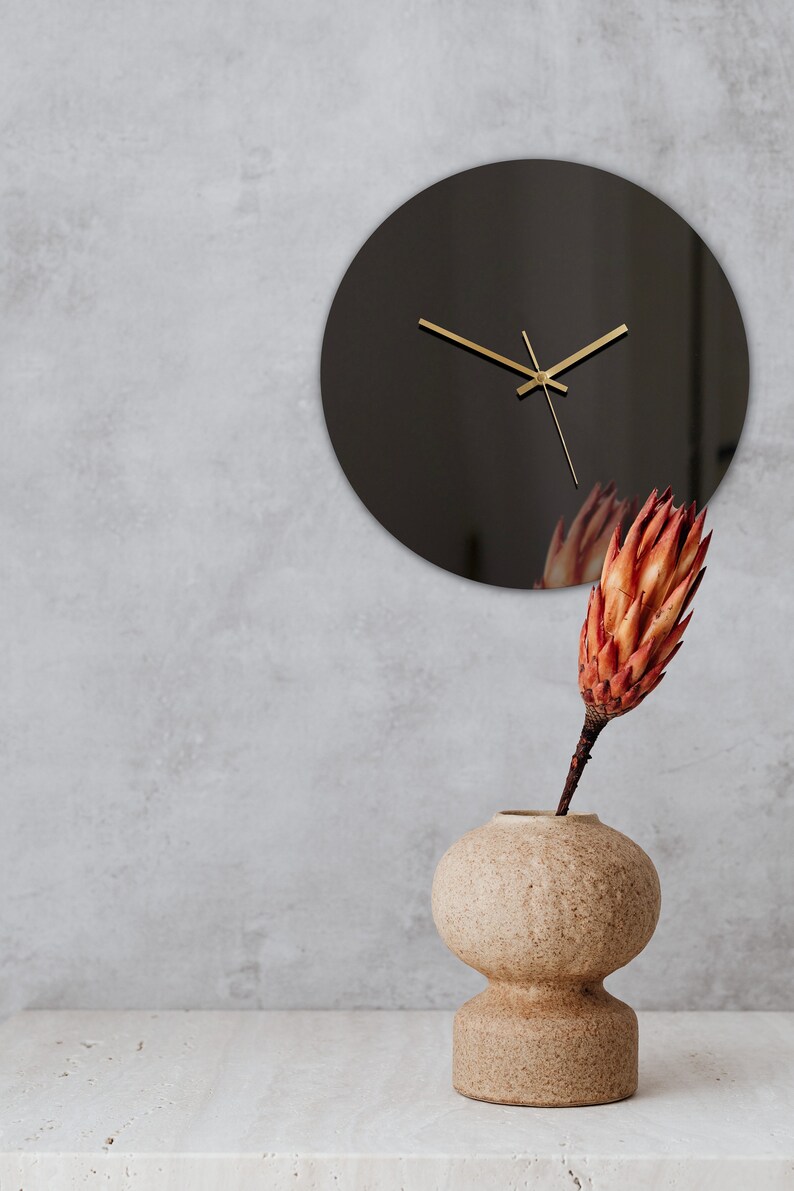 May include: A black round clock with gold hands hangs on a gray wall. A beige ceramic vase with a round body and a smaller round base holds a dried orange flower.