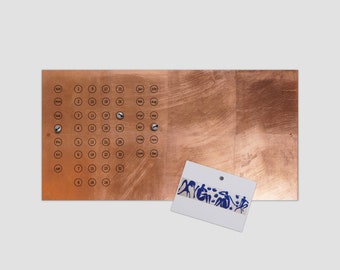 May include: A copper-colored calendar with a grid of numbered circles representing each day of the year. The calendar is mounted on a wall with screws. A white card with blue writing is attached to the calendar with a magnet.