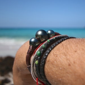 May include: A multi-strand bracelet featuring two large, dark, lustrous spherical pearls on a brown leather cord, accompanied by a thin red cord, a silver-toned metallic chain, and a black braided leather cord accented with small green beads.