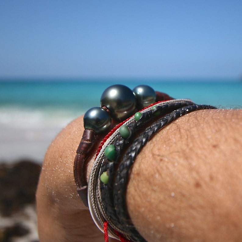 May include: A multi-strand bracelet featuring two large, dark, lustrous spherical pearls on a brown leather cord, accompanied by a thin red cord, a silver-toned metallic chain, and a black braided leather cord accented with small green beads.