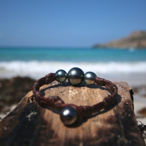 May include: A bracelet featuring three dark, lustrous spherical pearls centred on a braided brown leather cord, with a single matching pearl serving as a clasp closure. The design highlights the contrast between the smooth, dark pearls and the textured leather.