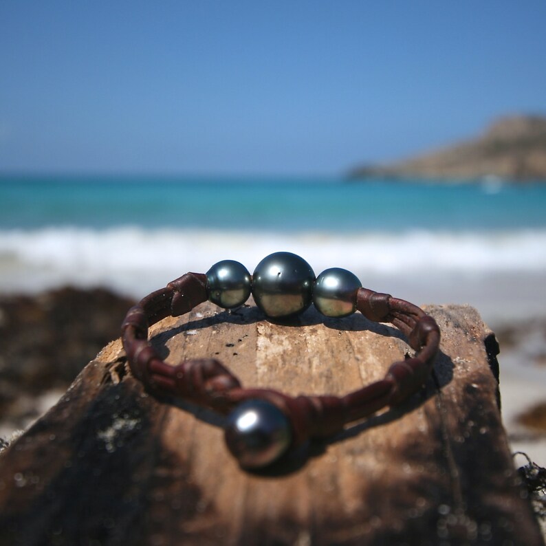 May include: A bracelet featuring three dark, lustrous spherical pearls centred on a braided brown leather cord, with a single matching pearl serving as a clasp closure. The design highlights the contrast between the smooth, dark pearls and the textured leather.
