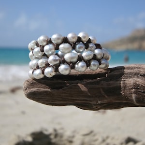 May include: A white pearl bracelet with brown leather cord wrapping around the pearls. The bracelet is on a piece of driftwood.