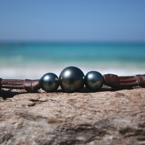 May include: A bracelet featuring three dark, lustrous, spherical pearls centred on a brown leather cord. The largest pearl is positioned in the middle, flanked by two smaller pearls of the same dark, reflective colour, secured by the leather band.