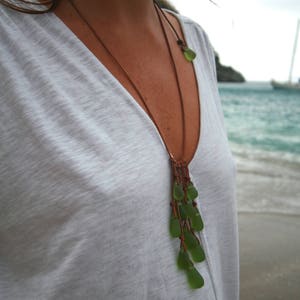 May include: A long necklace featuring multiple smooth, translucent green sea glass pieces suspended from a thin brown leather cord. The glass elements are arranged in a cascading cluster, with a single matching green sea glass bead accenting the cord.