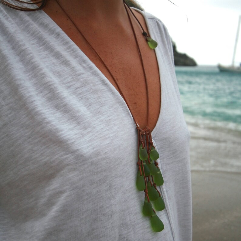 May include: A long necklace featuring multiple smooth, translucent green sea glass pieces suspended from a thin brown leather cord. The glass elements are arranged in a cascading cluster, with a single matching green sea glass bead accenting the cord.