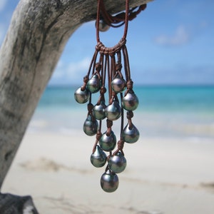 May include: A necklace featuring multiple dark, iridescent, teardrop-shaped pearls suspended from a brown leather cord. The pearls exhibit shades of charcoal, green, and blue, arranged in a cascading cluster design attached to the leather strands.