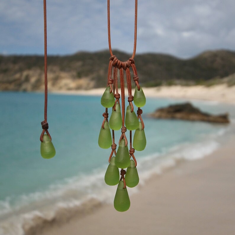 May include: A necklace featuring multiple light green, teardrop-shaped glass beads suspended from thin brown leather cords. The beads are arranged in a cascading cluster, with each piece individually knotted to the leather cord structure.