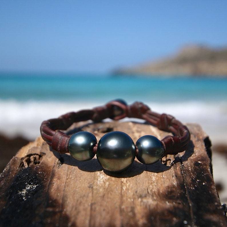 May include: A bracelet featuring three dark, lustrous spherical pearls centred on a braided brown leather cord. The central pearl is larger than the two flanking pearls, and the leather band is secured with a simple knot closure mechanism.