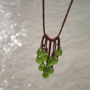 May include: A necklace featuring multiple teardrop-shaped, frosted light green glass beads suspended from a brown leather cord. The beads are arranged in a cluster, each attached to the cord with individual knots, creating a layered, dangling effect.