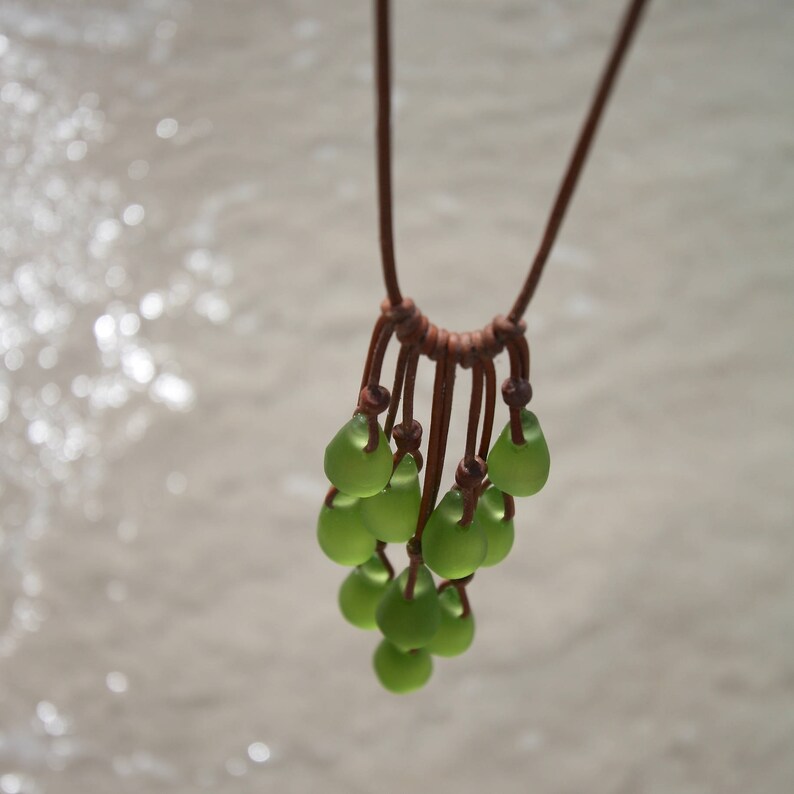 May include: A necklace featuring multiple teardrop-shaped, frosted light green glass beads suspended from a brown leather cord. The beads are arranged in a cluster, each attached to the cord with individual knots, creating a layered, dangling effect.