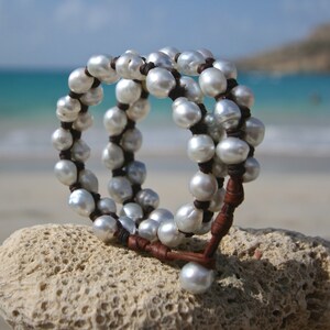May include: A triple-strand bracelet made of white pearls and brown leather. The bracelet is on a white coral-like surface.