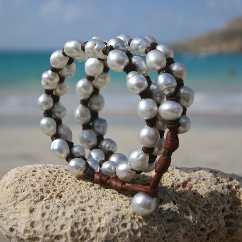 May include: A triple-strand bracelet made of white pearls and brown leather. The bracelet is on a white coral-like surface.