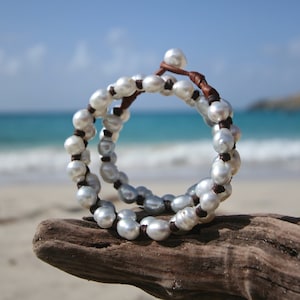 May include: A white pearl bracelet with brown leather cord, wrapped around a piece of driftwood on a beach.
