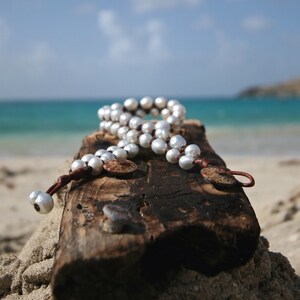 May include: A white pearl necklace with brown leather accents on a weathered piece of driftwood. The necklace is on a beach with blue water in the background.