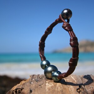 May include: A bracelet featuring a dark brown leather cord with a series of knots securing four dark, iridescent, spherical pearls. The pearls vary slightly in size, with one positioned at the clasp and three clustered together along the band.