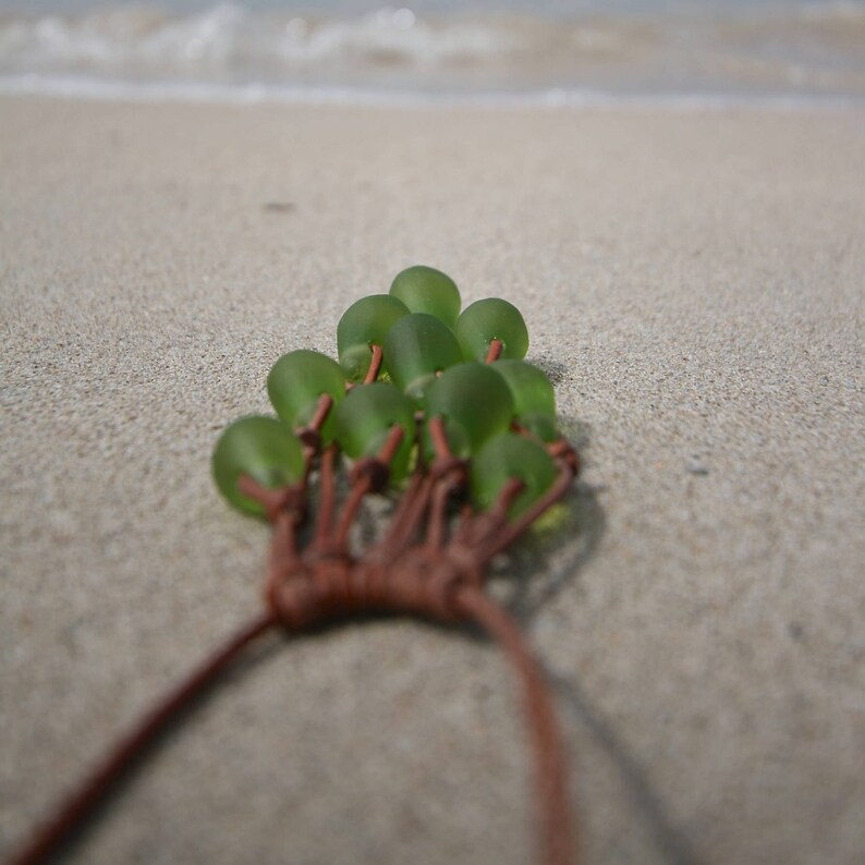 May include: A necklace featuring multiple smooth, frosted green sea glass beads arranged in a cluster. The beads are connected by thin, brown leather cords that are knotted together at the base, extending into two long strands for fastening.