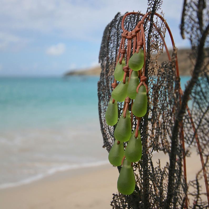 May include: A cluster of ten smooth, translucent, light green sea glass pieces shaped like teardrops. Each piece is individually suspended by a thin, tan leather cord, creating a cascading arrangement that hangs from a dark, fan-shaped coral structure.