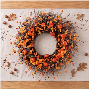 Può includere: Una ghirlanda circolare realizzata con ramoscelli marrone scuro, decorata con piccole zucche arancioni, foglie arancioni vivaci e minuscole bacche arancioni. La composizione presenta un design denso e strutturato con steli e rami sporgenti.