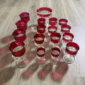 Puede incluir: Una colección de copas de cristal transparente y un cuenco con bordes de color magenta intenso. El conjunto incluye varios tamaños de vasos con bases circulares y patrones de vidrio texturizado, dispuestos sobre una superficie de madera clara.