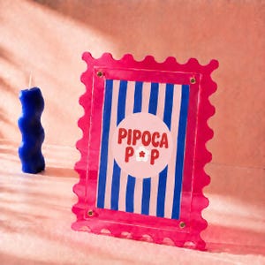 Peut inclure: Un cadre rectangulaire en acrylique rose avec un bord festonné, présentant une impression centrale à rayures verticales bleues et blanches et une étiquette ronde blanche affichant le texte rouge PIPOCA POP avec une petite fleur blanche.