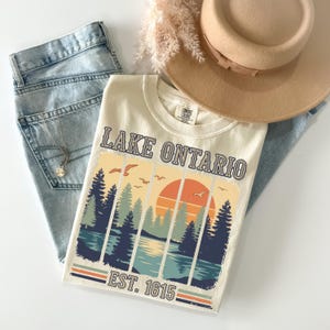 May include: A cream-colored t-shirt featuring a graphic of Lake Ontario with a sunset, pine trees, and birds. The text reads Lake Ontario and Est. 1615. The shirt is paired with light blue denim jeans and a tan wide-brimmed hat with a ribbon band.