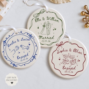May include: Three round white ceramic ornaments with white ribbon hangers. One features blue ink with hands and an engagement ring, one green ink with a couple, and one maroon ink with champagne glasses. Each has names, dates, and decorative borders.