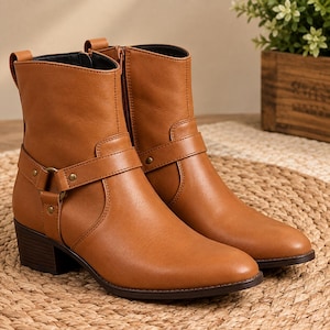 Könnte beinhalten: Ein Paar hellbraune Stiefeletten mit glatter Oberfläche, einem niedrigen Blockabsatz und einem dekorativen Riemen über dem Knöchel, der mit einem Metallring und Nieten befestigt ist. Die Stiefel haben einen seitlichen Reißverschluss und eine Zuglasche.