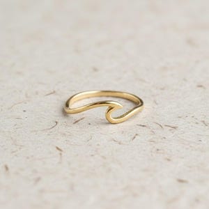 Könnte beinhalten: Ein goldfarbener Ring mit einem durchgehenden Band, das sich an der Oberseite zu einer stilisierten Wellenform krümmt. Das Metall hat eine glatte, polierte Oberfläche und das Design besteht aus einem einzigen Stück Metall, das den Ring und die Welle bildet.