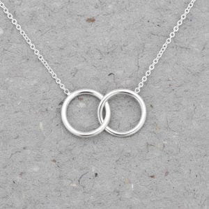May include: A silver-toned necklace featuring two interlocking circular rings as the central pendant. The pendant is suspended from a delicate, thin silver-toned chain with small, uniform links that extend outwards from each side of the circles.