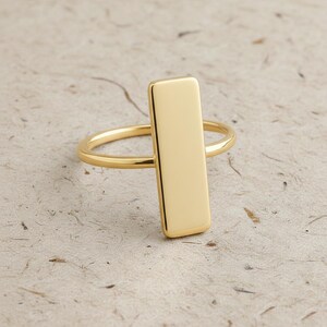 May include: A gold-toned ring featuring a smooth, polished vertical rectangular bar centred on a thin, rounded band. The metal surface is reflective and uniform in colour, showcasing a clean, geometric design with rounded edges on the bar.