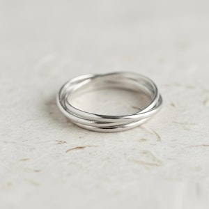 Può includere: Un anello color argento composto da tre sottili fasce intrecciate che si sovrappongono per creare una forma circolare continua. La superficie metallica presenta una finitura liscia e lucida che riflette la luce, evidenziando il profilo.