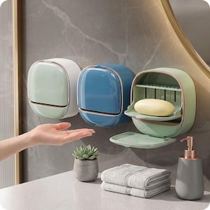 Può includere: Tre portasapone da parete in verde chiaro, blu e verde salvia con coperchi a cerniera. Il supporto a destra è aperto, rivelando una saponetta gialla all'interno. Un dispenser di sapone grigio, una piccola pianta grassa e un asciugamano grigio piegato.