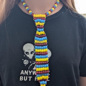 May include: A necktie-shaped necklace crafted from plastic pony beads in alternating horizontal rows of yellow, light blue, and dark red. The beads are strung together with thin neon orange cord, forming a structured tie shape suspended from a beaded chain.