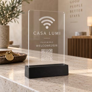 Può includere: Un cartello rettangolare in acrilico trasparente su una base rettangolare nera. Il cartello presenta un simbolo Wi-Fi bianco, il testo CASA LUMI, la parola PASSWORD, il testo WELCOME2026 e un codice QR quadrato sulla superficie.