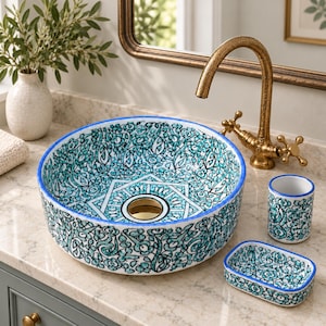 Könnte beinhalten: Ein rundes Aufsatzwaschbecken aus Keramik mit einem blaugrünen und weißen Blumenmuster, einem geometrischen Design in der Mitte und einem goldenen Abfluss. Das Set enthält einen passenden zylindrischen Zahnputzbecher und eine rechteckige Seifenschale.