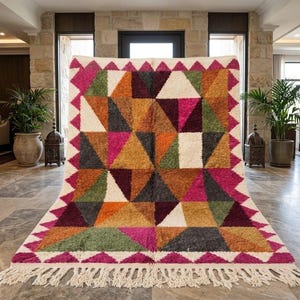 Puede incluir: Una alfombra rectangular con un patrón geométrico de triángulos en tonos magenta, verde oliva, amarillo mostaza, marrón oscuro y crema. La alfombra tiene un pelo grueso y texturizado y un borde de triángulos magenta con flecos blancos.