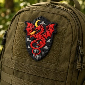 Puede incluir: Una mochila táctica de color verde oliva con un parche de PVC en forma de escudo. El parche muestra un dragón rojo escupiendo fuego, una luna creciente amarilla y nubes grises sobre la silueta de una catedral gótica en negro.