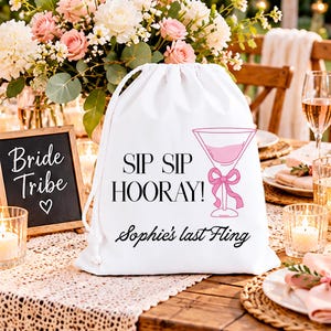 Puede incluir: Una bolsa de cordón blanca con el texto negro SIP SIP HOORAY! y Sophie's last Fling, acompañada de un gráfico rosa de una copa de martini con un lazo. La bolsa está hecha de tela e incluye cordones blancos para el cierre en la parte superior.
