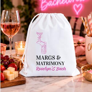 May include: A white drawstring bag featuring a pink graphic of a cowboy boot holding a martini glass, with the black text MARGS & MATRIMONY and the pink script text Roselyn's Bach printed on the front of the fabric pouch.