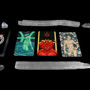 May include: Three tarot cards on a black background with crystals and selenite wands. The cards are the Queen of Pisces, the Devil, and the Emperor. The Queen of Pisces card features a woman with green skin and long hair, sitting in a pool of water. The Devil card features a red demon with three heads and horns. The Emperor card features a man with a sword, standing in front of a pink and white background.