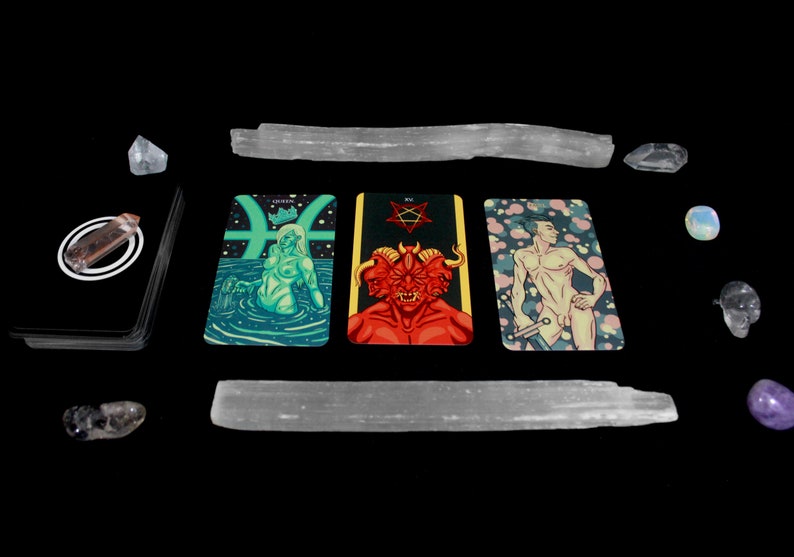 May include: Three tarot cards on a black background with crystals and selenite wands. The cards are the Queen of Pisces, the Devil, and the Emperor. The Queen of Pisces card features a woman with green skin and long hair, sitting in a pool of water. The Devil card features a red demon with three heads and horns. The Emperor card features a man with a sword, standing in front of a pink and white background.